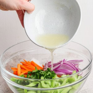 Person pouring a light dressing into a bowl of chopped vegetables including carrots, red onions, celery, and herbs.