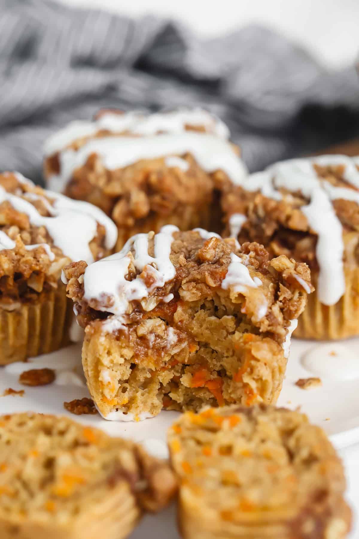 Carrot muffins with crumble topping and white icing drizzled on top, one muffin partially eaten, displayed on a white surface.