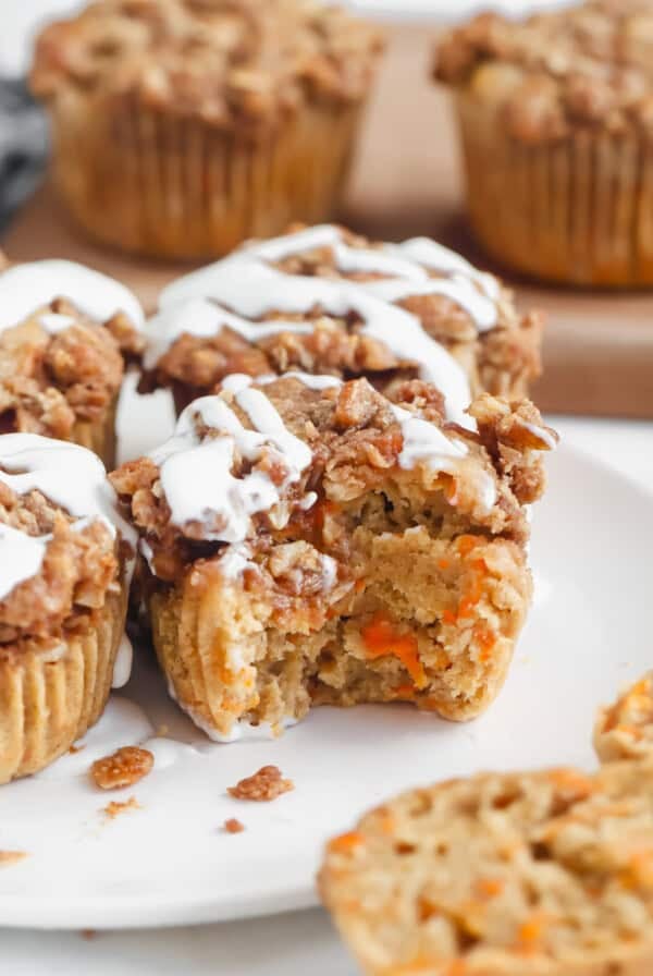 Muffins with a crumb topping and white icing drizzle on a plate, one muffin partially eaten, displaying an orange and beige interior.