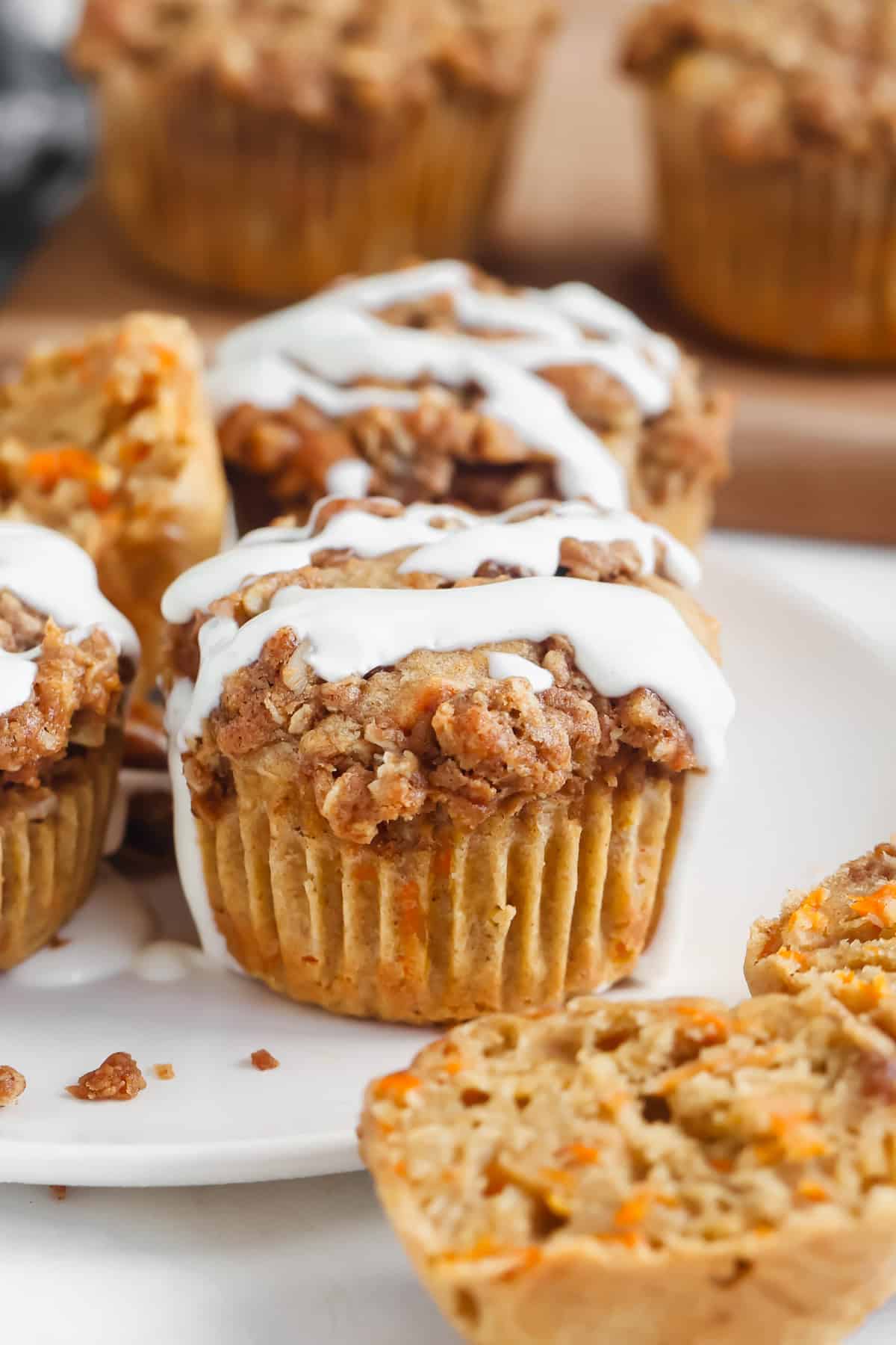 Carrot muffins with streusel topping and white icing are displayed on a plate, with one muffin cut in half.