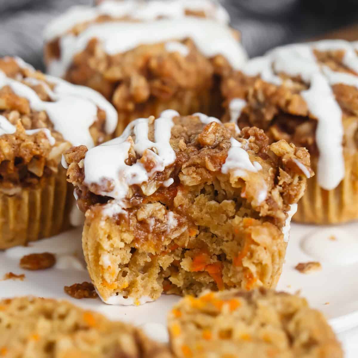 Close-up of a carrot muffin with a crumbly topping and white icing. One muffin has a bite taken out, revealing a moist interior with visible carrot pieces.