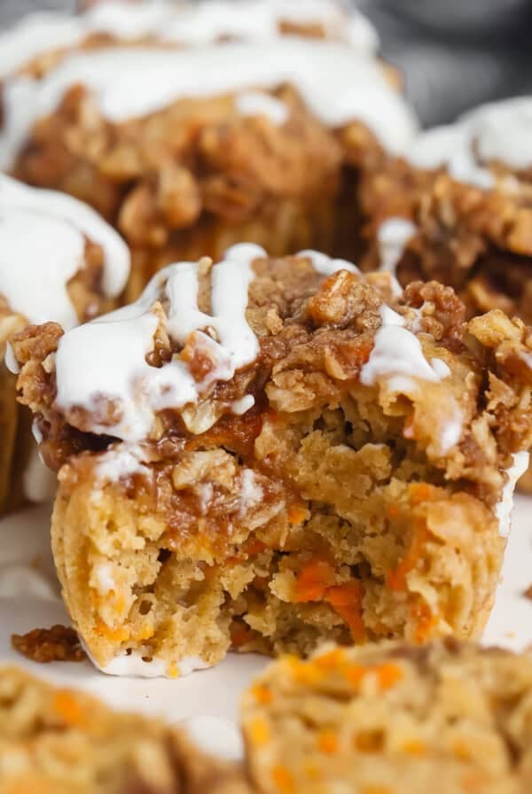 Close-up of a carrot muffin with a crumbly topping and white icing. One muffin has a bite taken out, revealing a moist interior with visible carrot pieces.