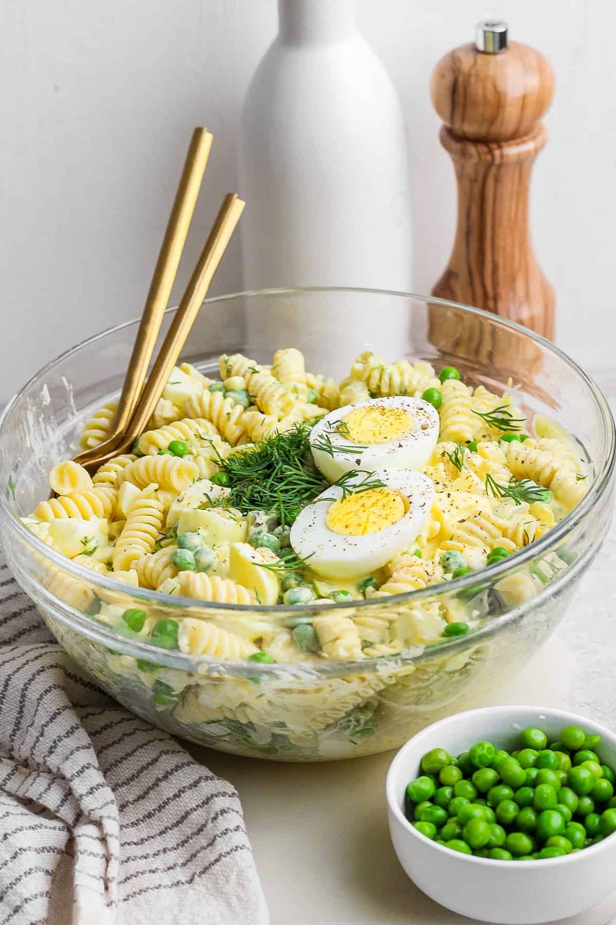 Glass bowl of pasta salad with sliced boiled eggs and herbs, garnished with peas. Placed on a table with chopsticks, a white towel, a small bowl of peas, a pepper mill, and an olive oil bottle.