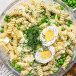 A bowl of fusilli pasta salad with green peas, chopped dill, and sliced hard-boiled egg halves on top.