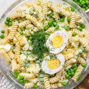A bowl of fusilli pasta salad with green peas, chopped dill, and sliced hard-boiled egg halves on top.