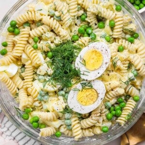 A bowl of pasta salad with rotini, green peas, hard-boiled egg slices, dill, and a creamy dressing. A gold spoon and a cloth napkin are next to the bowl.