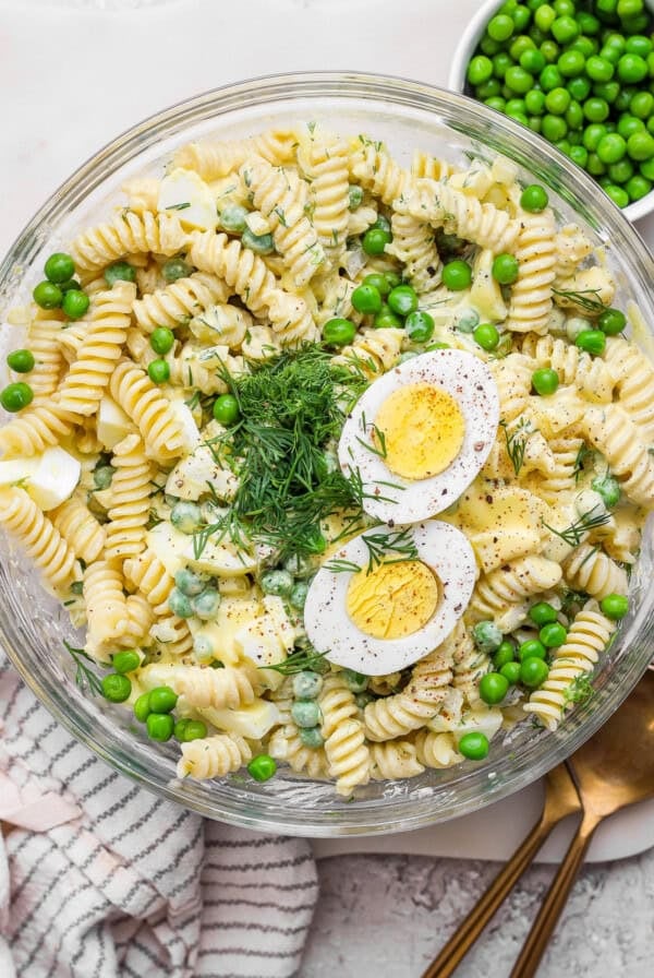 A bowl of pasta salad with rotini, green peas, hard-boiled egg slices, dill, and a creamy dressing. A gold spoon and a cloth napkin are next to the bowl.