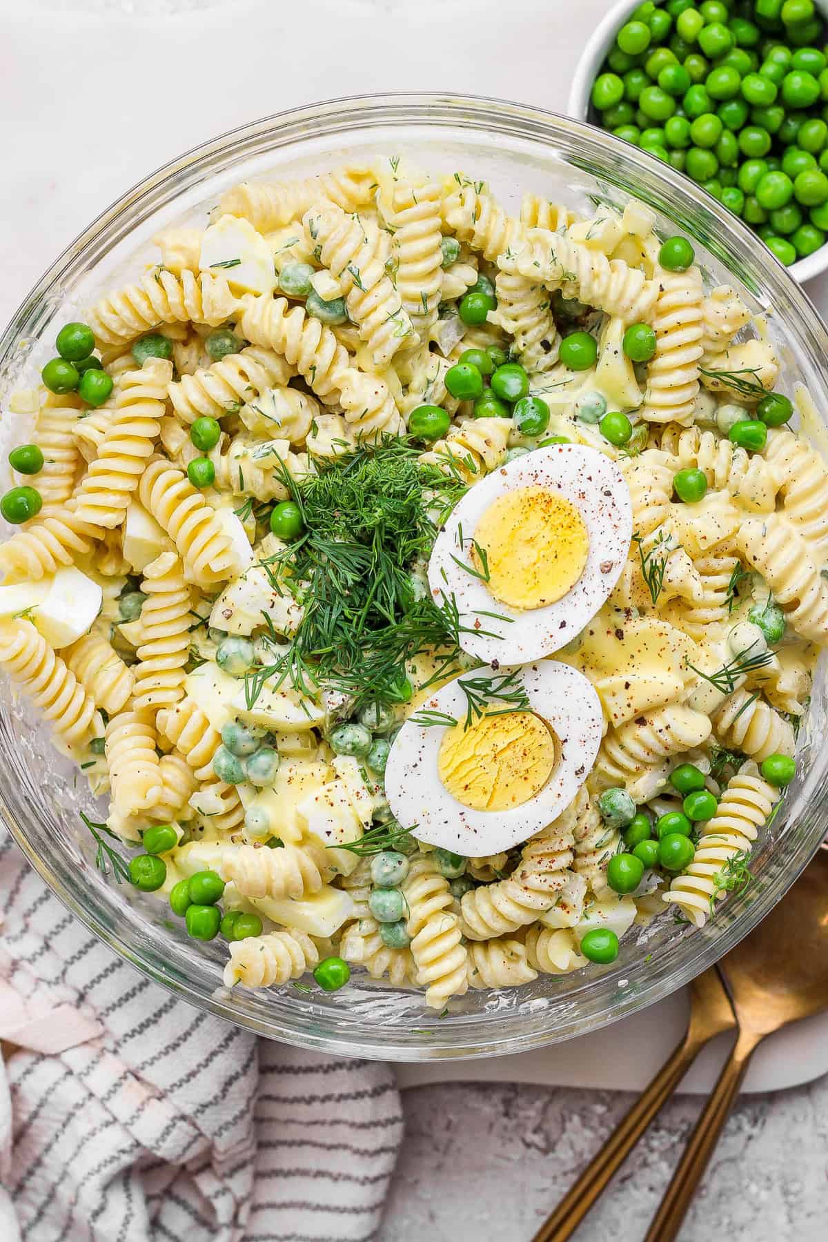 A bowl of pasta salad with rotini, green peas, hard-boiled egg slices, dill, and a creamy dressing. A gold spoon and a cloth napkin are next to the bowl.