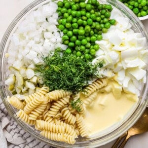 Glass bowl containing rotini pasta, peas, chopped onions, boiled eggs, pickles, fresh dill, and mayonnaise, set on a white surface with a silver spoon nearby.