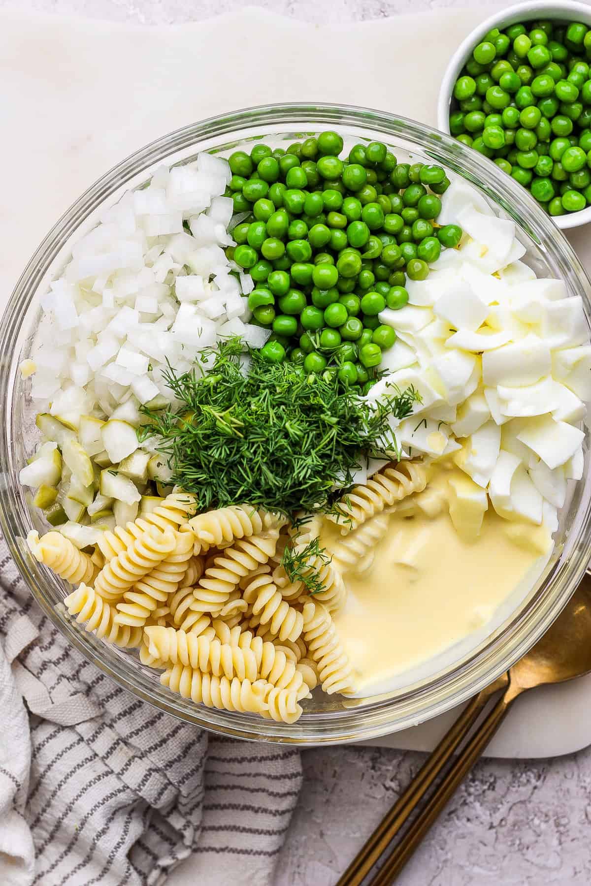 Glass bowl containing rotini pasta, peas, chopped onions, boiled eggs, pickles, fresh dill, and mayonnaise, set on a white surface with a silver spoon nearby.