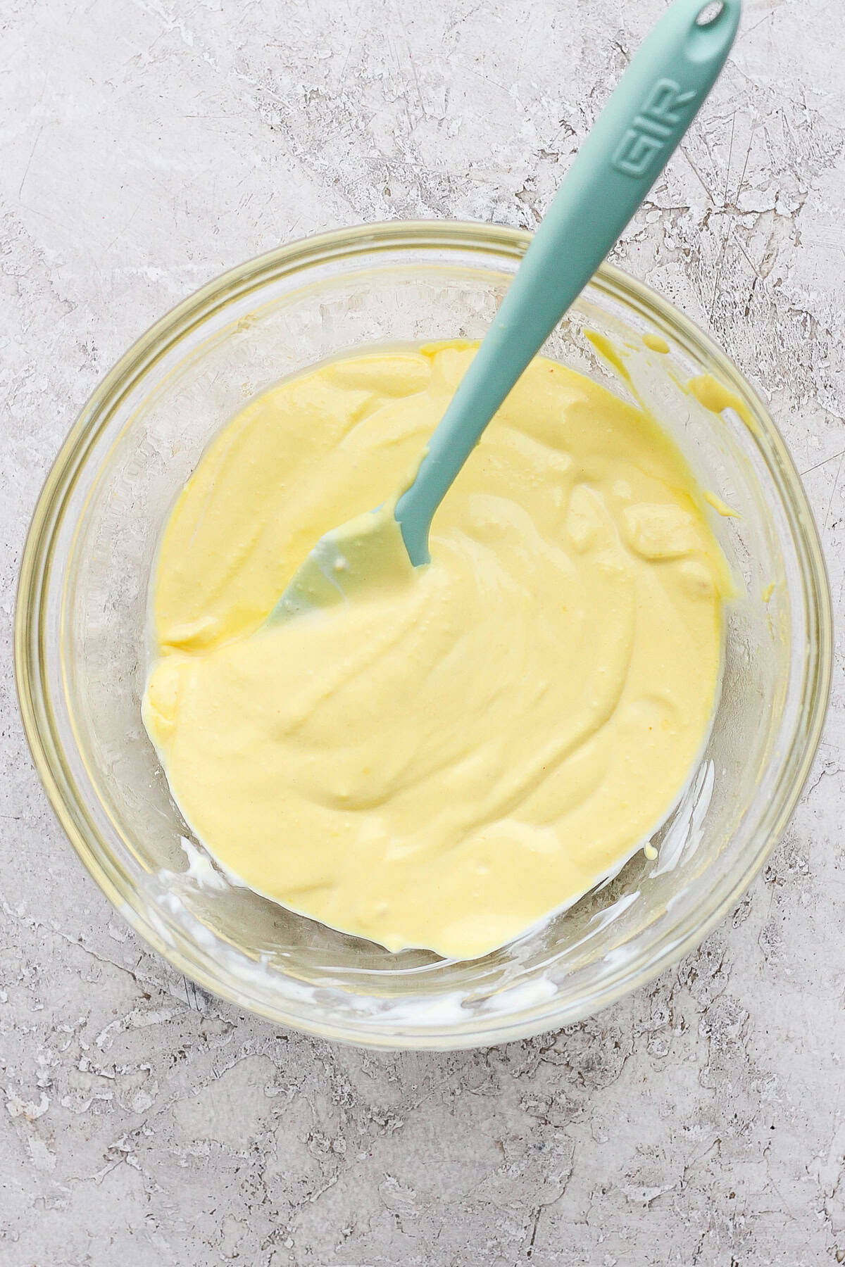 A glass bowl containing creamy, yellow batter with a teal spatula on a textured gray surface.