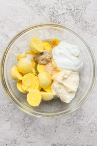 Bowl containing boiled egg yolks, mayonnaise, mustard, yogurt, and seasoning on a textured surface.