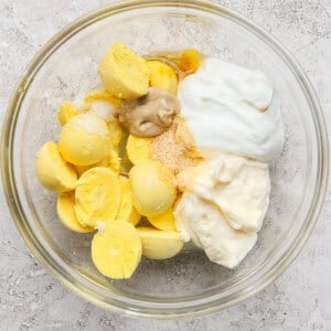 Bowl containing boiled egg yolks, mayonnaise, mustard, yogurt, and seasoning on a textured surface.