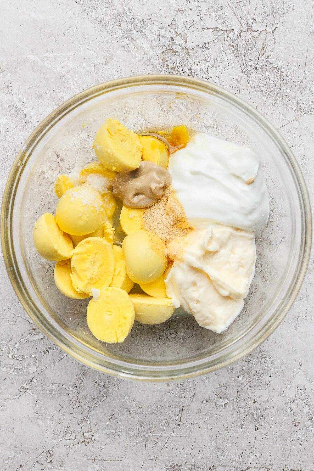 Bowl containing boiled egg yolks, mayonnaise, mustard, yogurt, and seasoning on a textured surface.