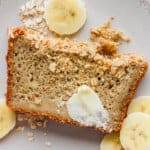 Slice of banana bread with butter and oats, surrounded by banana slices, on a white plate.