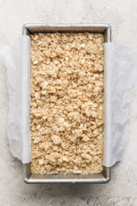 A rectangular baking dish lined with parchment paper contains an unbaked oat crumble mixture on a light textured surface.