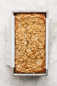 Loaf of banana bread with oat crumble topping in a parchment-lined metal baking pan on a light textured surface.