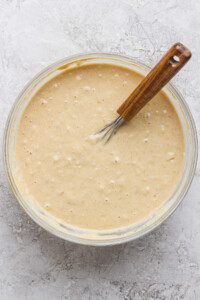 A glass bowl filled with thick, creamy batter is placed on a textured gray surface. A wooden-handled whisk rests inside the bowl.