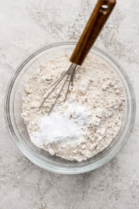A glass bowl contains a mixture of flour and baking powder, with a metal whisk resting inside, placed on a textured gray surface.