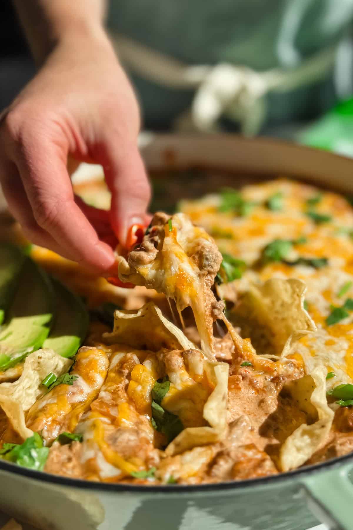 Hand reaching into a dish of cheesy nachos topped with cilantro and sliced avocado.