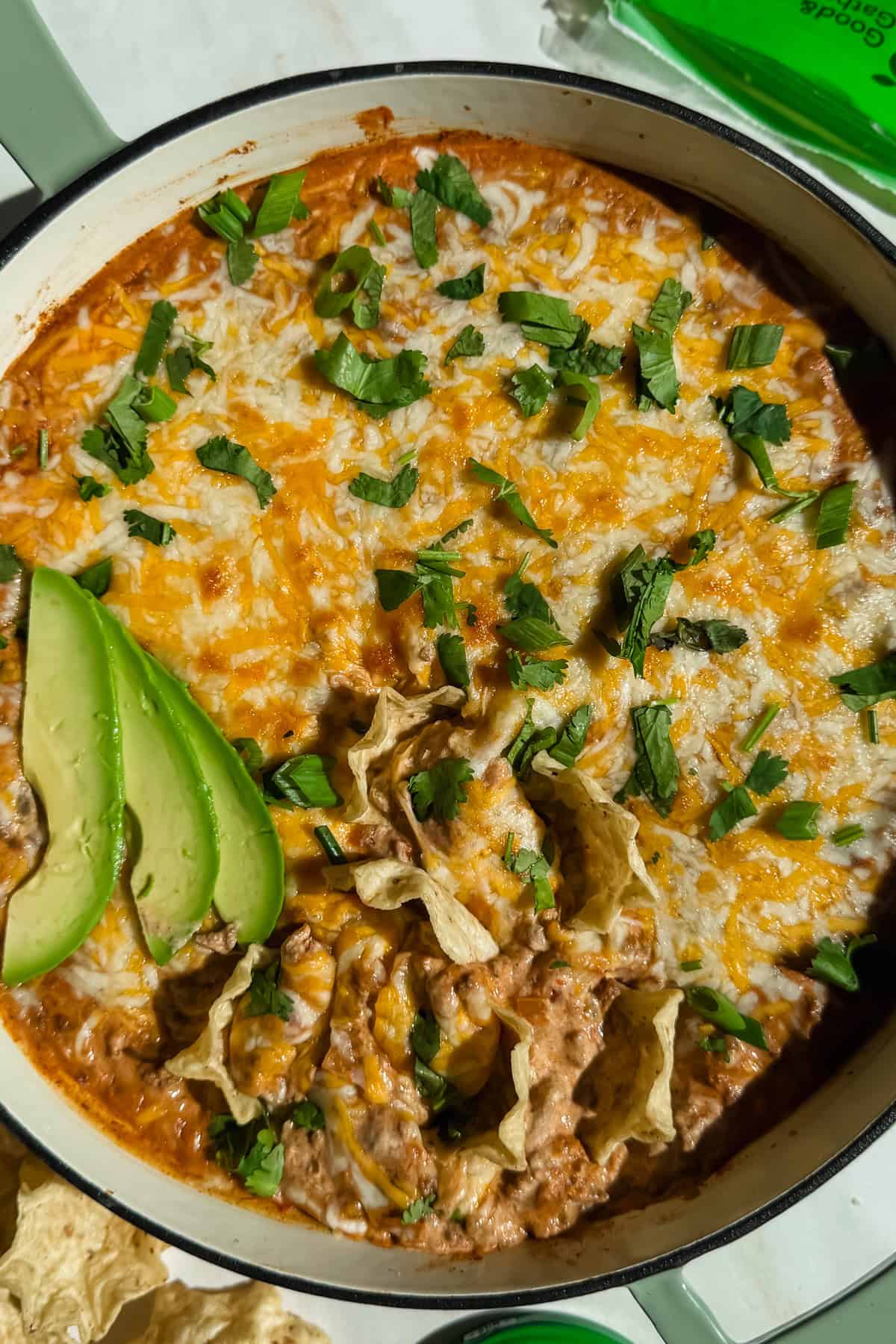 A dish of cheesy dip topped with cilantro and avocado slices in a white casserole dish, surrounded by tortilla chips.