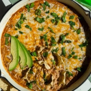 A baked dish topped with melted cheese, garnished with sliced avocado and fresh herbs, next to tortilla chips.