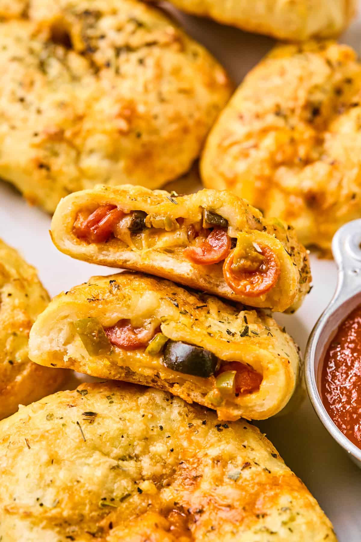 Close-up of baked calzones, reminiscent of pizza hot pockets, filled with cheese, pepperoni, and vegetables, accompanied by a small dish of marinara sauce.