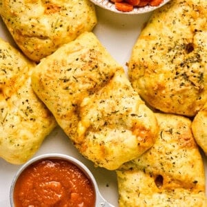 Golden-brown stuffed bread rolls, reminiscent of delightful pizza hot pockets, are sprinkled with herbs and paired with a small bowl of tomato sauce on a white surface.