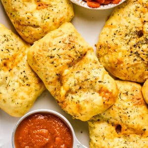 Golden, herb-seasoned bread parcels resembling mini pizza hot pockets sit beside a small dish of sliced pepperoni and a cup of marinara sauce.