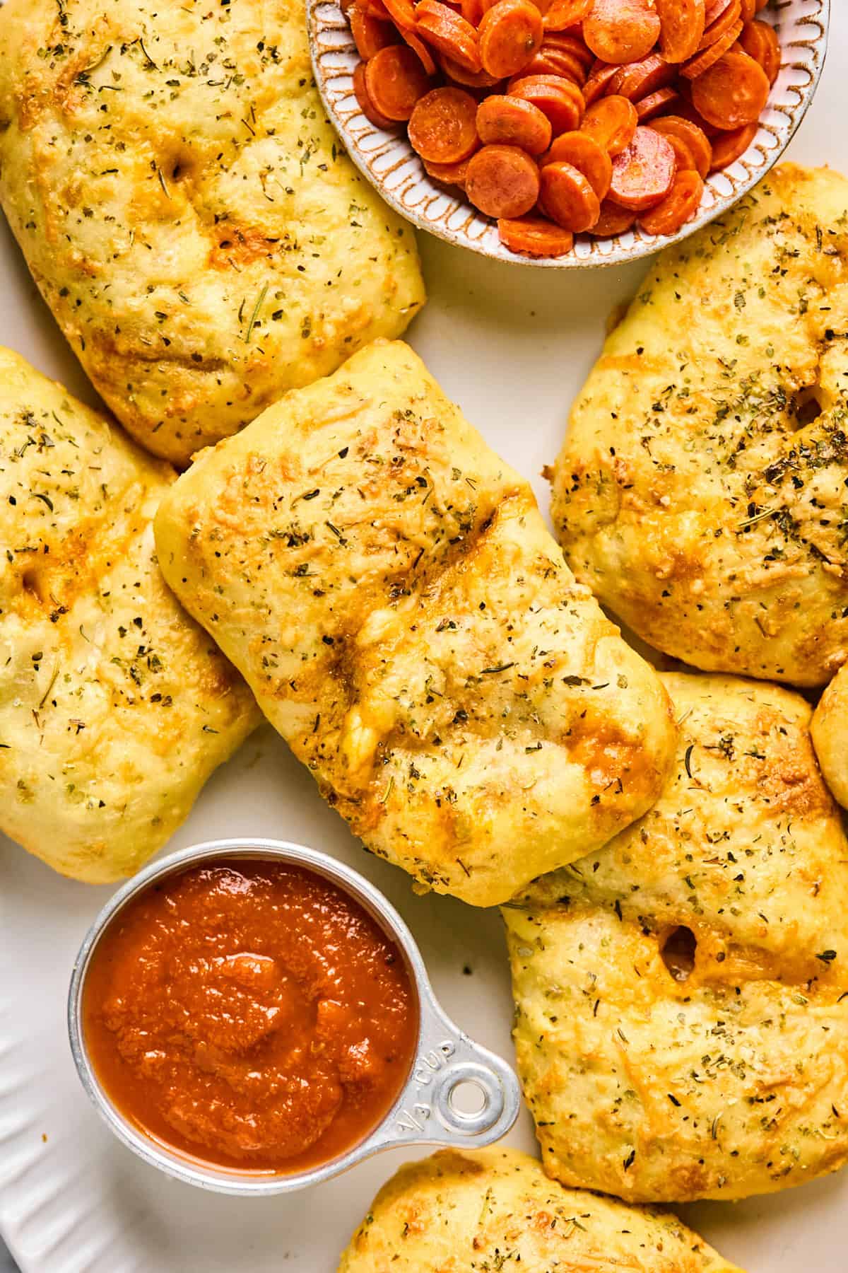 Golden, herb-seasoned bread parcels resembling mini pizza hot pockets sit beside a small dish of sliced pepperoni and a cup of marinara sauce.