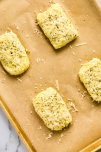 Four rectangular pizza hot pockets of seasoned dough rest on a parchment-lined baking sheet, sprinkled with herbs and cheese.