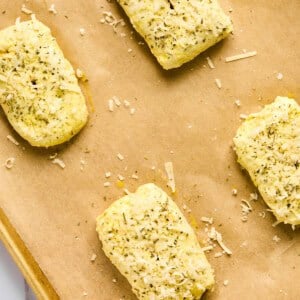 Four rectangular pizza hot pockets of seasoned dough rest on a parchment-lined baking sheet, sprinkled with herbs and cheese.