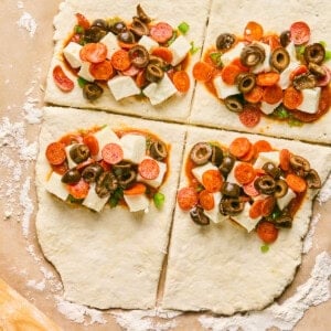 Four sections of raw dough, resembling pizza hot pockets, are topped with cheese cubes, pepperoni slices, olives, and green onions on a floured surface.