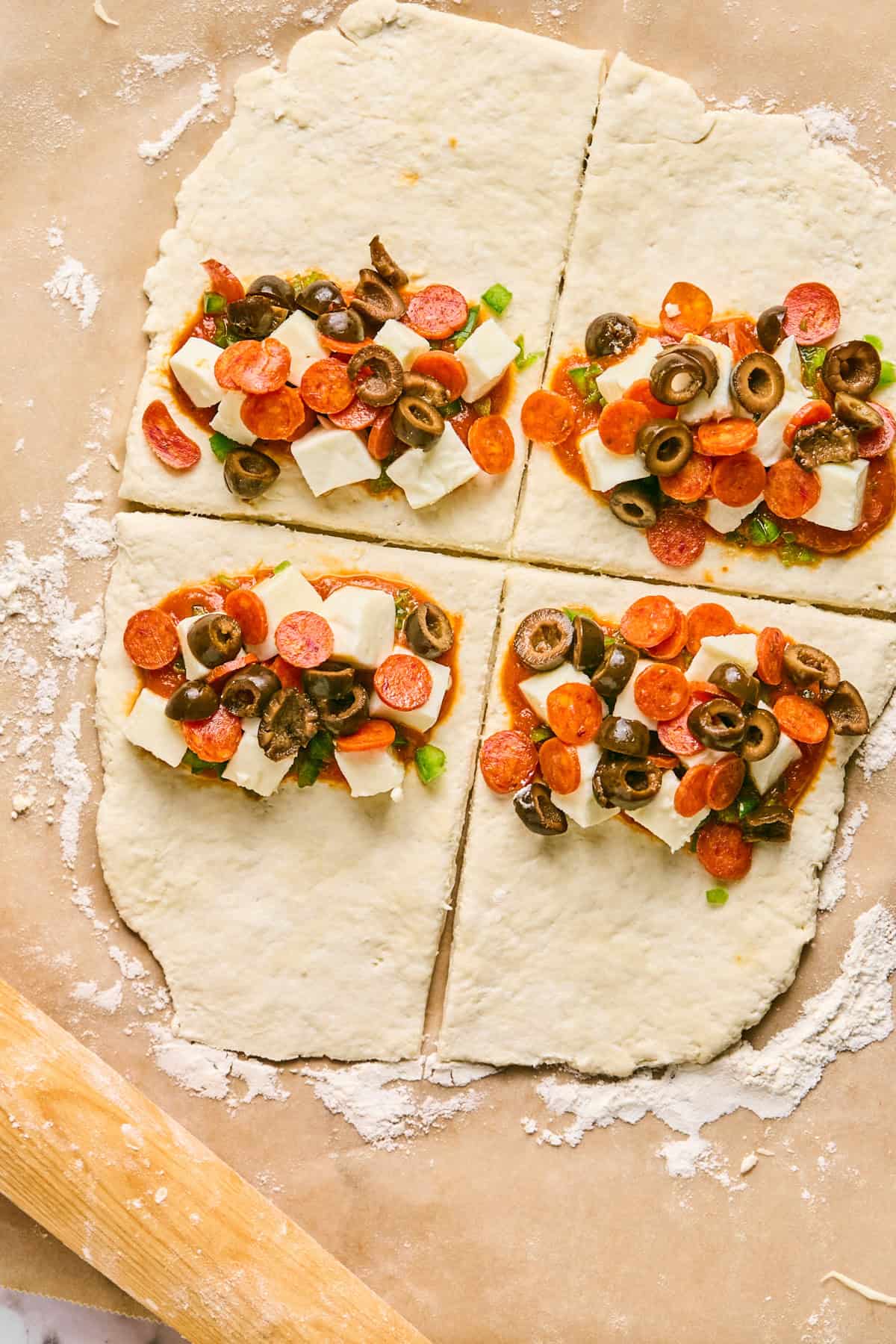 Four sections of raw dough, resembling pizza hot pockets, are topped with cheese cubes, pepperoni slices, olives, and green onions on a floured surface.