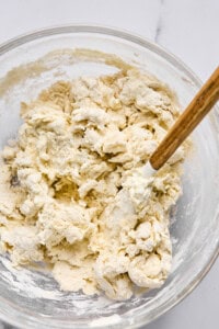 A glass bowl containing partially mixed dough with a wooden spoon sits ready to become delicious homemade pizza or hot pockets.