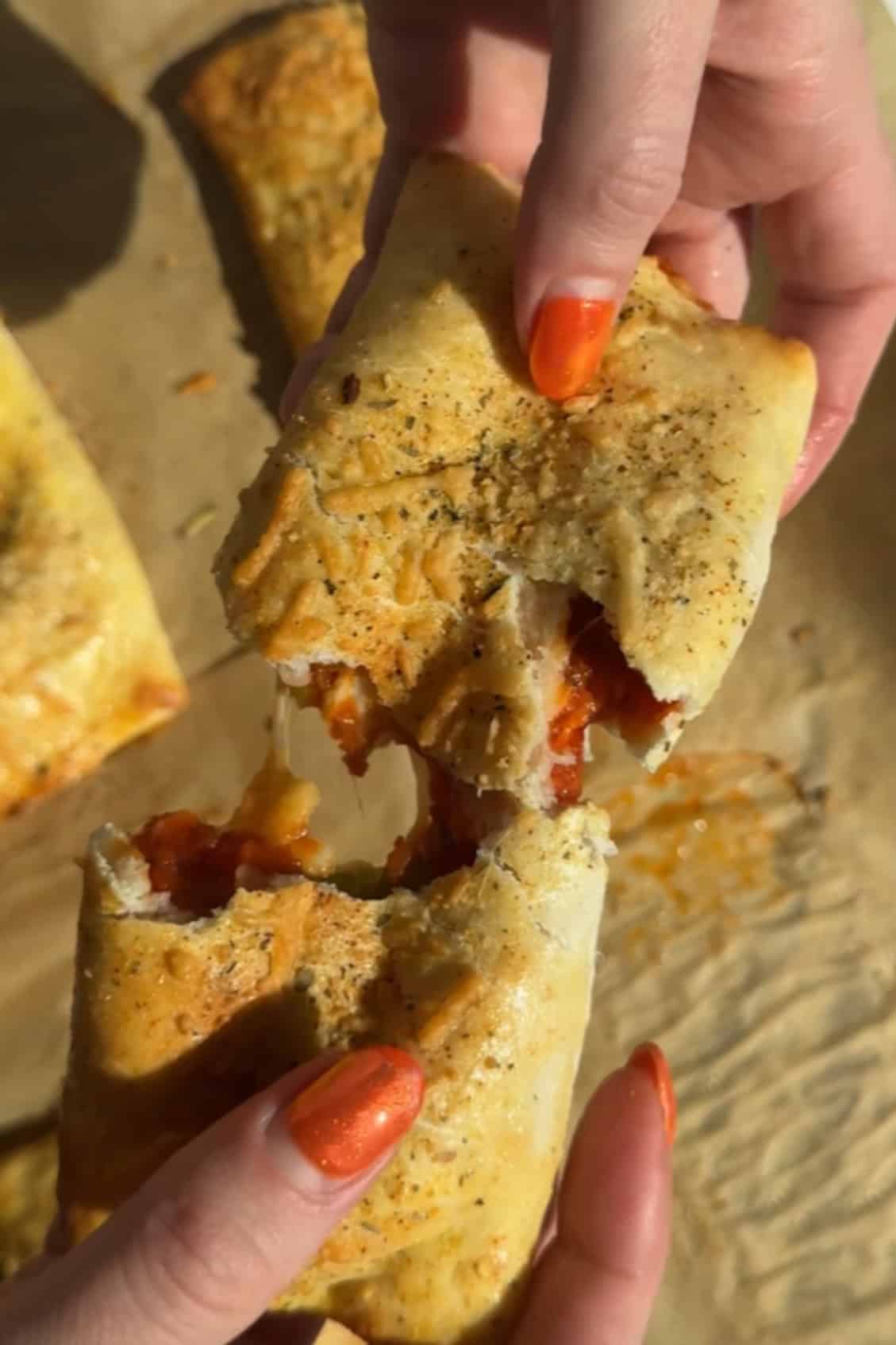 Hands tearing a baked calzone, reminiscent of tasty pizza pockets, revealing melted cheese and tomato sauce inside. The calzone is nestled on parchment paper.