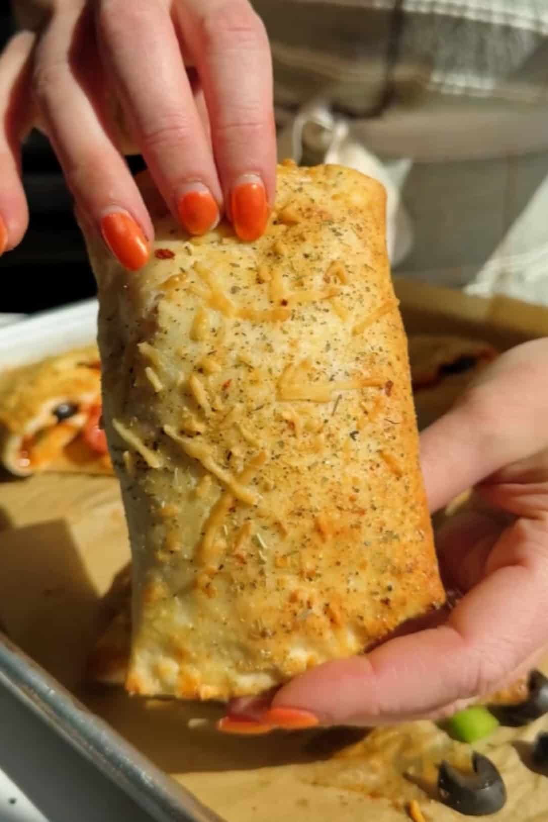 A person holds a baked calzone with a golden crust on a parchment-lined baking sheet, reminiscent of pizza hot pockets.