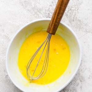 Whisk in a bowl of beaten eggs on a textured, light-colored surface.
