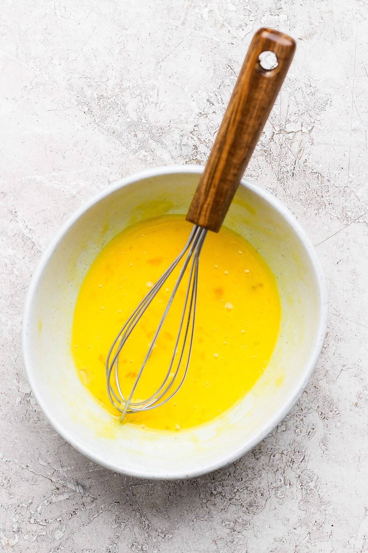 Whisk in a bowl of beaten eggs on a textured, light-colored surface.