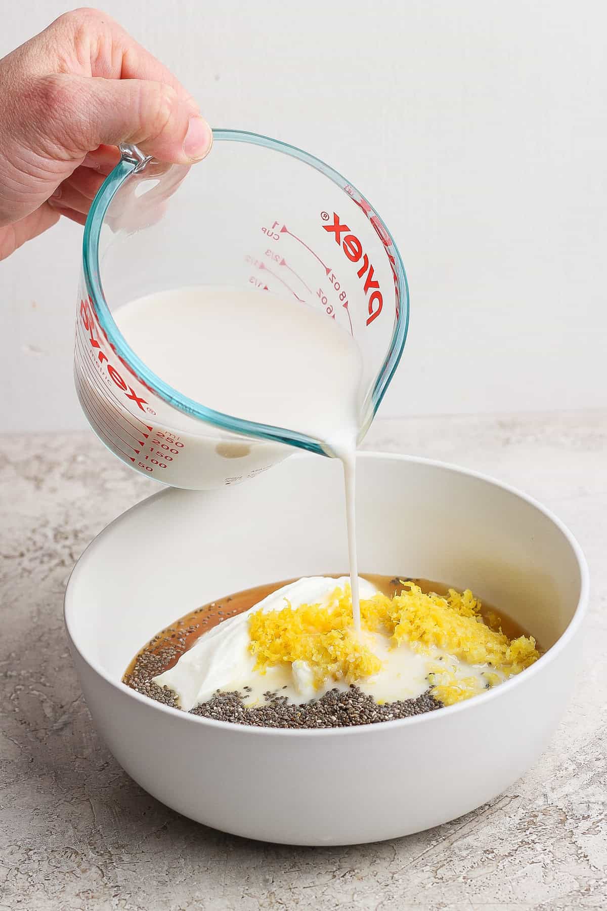 Pouring milk from a measuring cup into a bowl containing yogurt, chia seeds, lemon zest, and liquid.