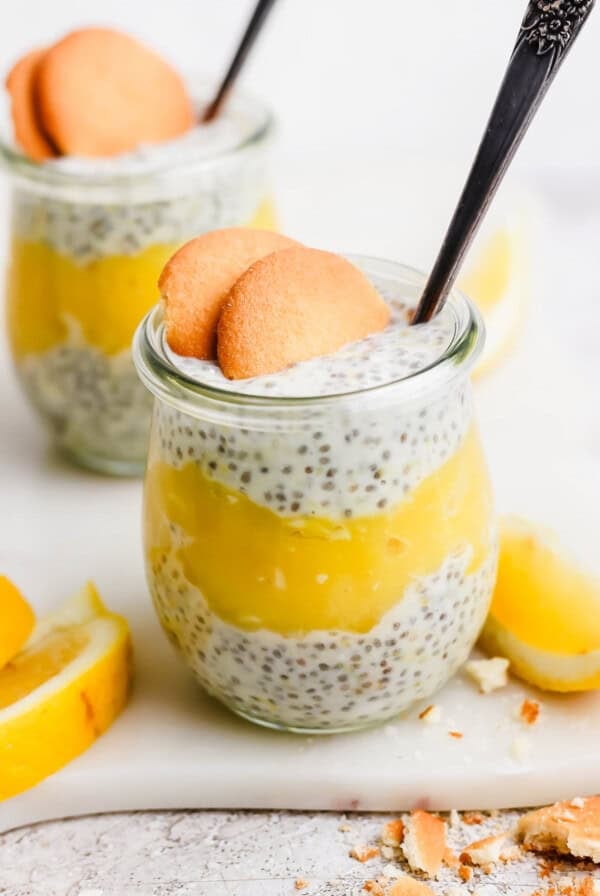 Two jars of chia pudding layered with yellow lemon curd, topped with two cookies and a spoon, surrounded by lemon slices.