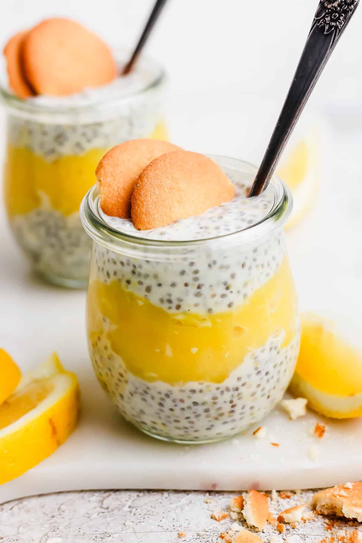 Two jars of chia pudding layered with yellow lemon curd, topped with two cookies and a spoon, surrounded by lemon slices.