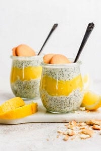 Two jars of chia pudding layered with yellow fruit puree, topped with two cookies each, on a white surface with lemon slices nearby.