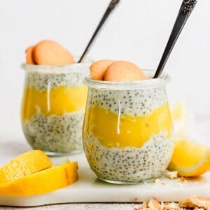 Two jars of chia pudding layered with yellow fruit puree, topped with two cookies each, on a white surface with lemon slices nearby.