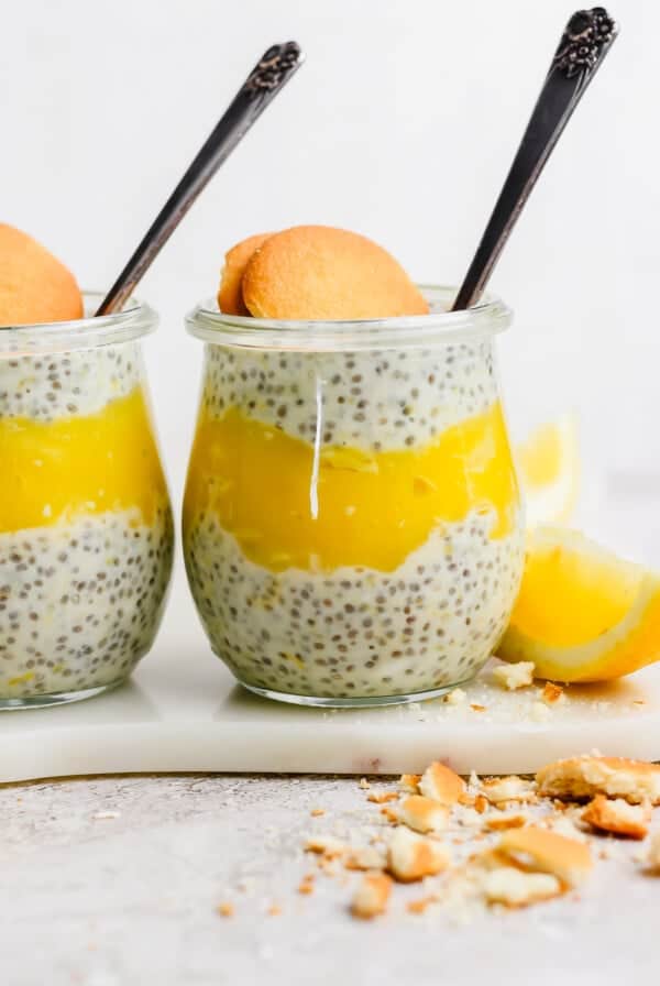 Two jars of chia pudding layered with lemon curd, each garnished with a cookie, placed on a marble surface with lemon wedges and cookie crumbs nearby.