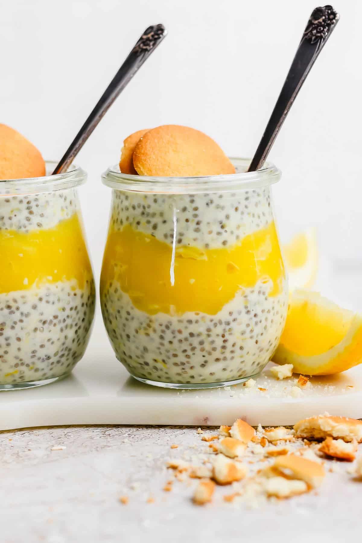 Two jars of chia pudding layered with lemon curd, each garnished with a cookie, placed on a marble surface with lemon wedges and cookie crumbs nearby.