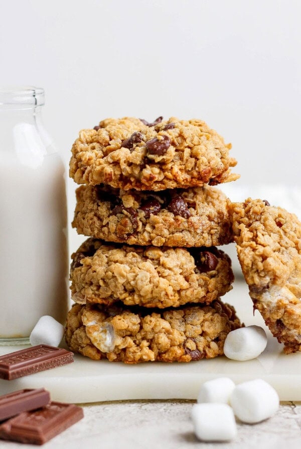 Stack of oatmeal chocolate chip cookies with scattered marshmallows, chocolate pieces, and a bottle of milk.