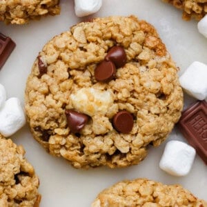 Close-up of an oatmeal cookie with chocolate chips and a marshmallow piece, surrounded by mini marshmallows and chocolate squares.