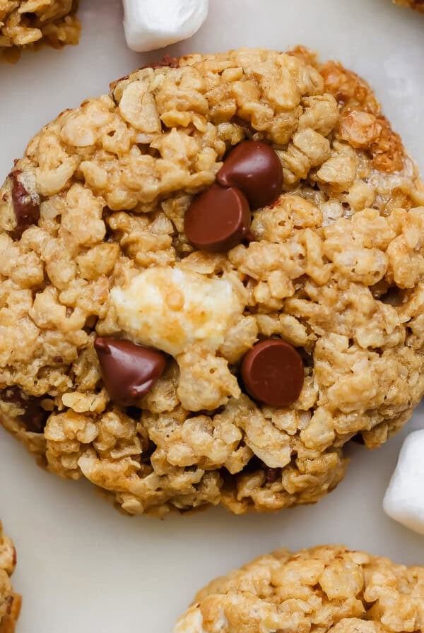 Close-up of an oatmeal cookie with chocolate chips and a marshmallow piece, surrounded by mini marshmallows and chocolate squares.