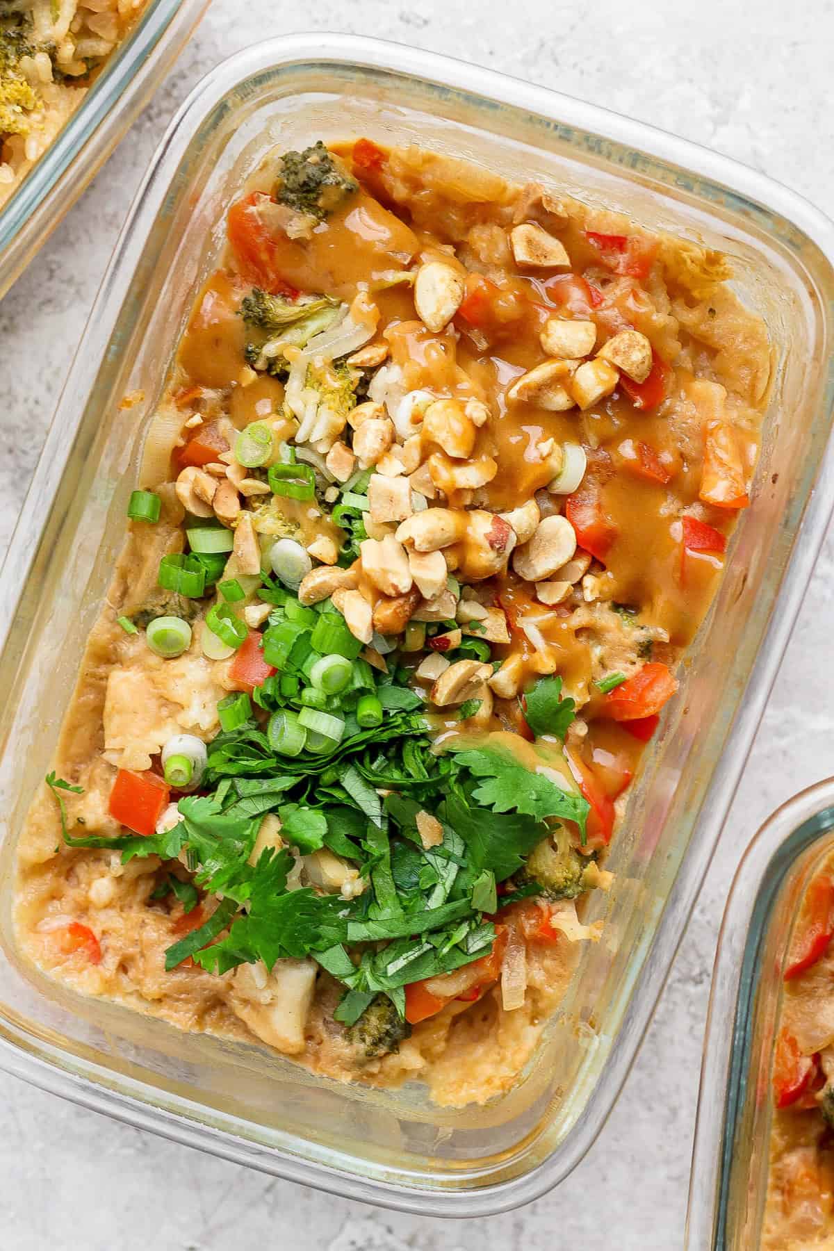 A glass container holds a colorful salad topped with chopped peanuts, green onions, and cilantro, accompanied by tender slices of peanut chicken, resting elegantly on a light textured surface.